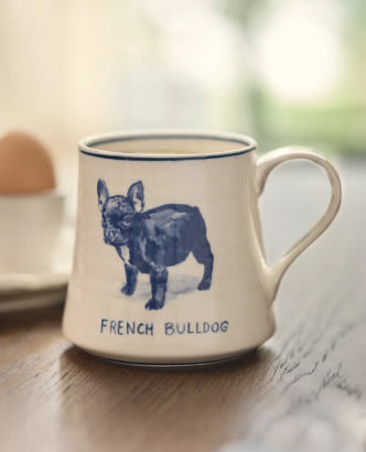 White ceramic mug with blue French Bulldog illustration and text "FRENCH BULLDOG" on wooden surface with blurred background.