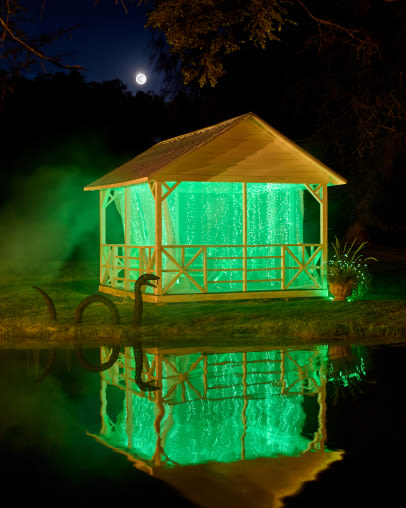 Illuminated gazebo with green lights reflecting in pond at night under full moon with trees silhouetted against dark blue sky.