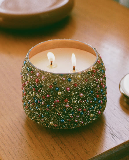 Two-wick candle in a decorative green vessel embellished with colorful beads and glitter, sitting on a wooden surface.