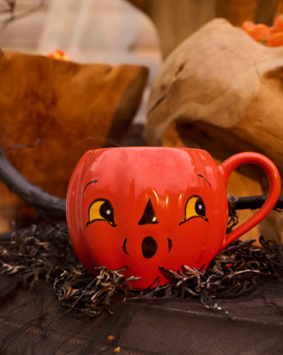 Orange pumpkin-shaped mug with carved jack-o-lantern face sits on dark surface with autumn decorations and paper bags in background.