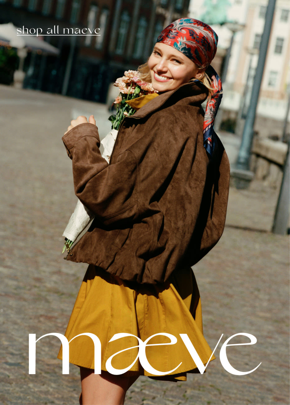 Smiling woman in colorful headscarf, brown jacket, and yellow skirt holding flowers outdoors. Text reads shop all maeve.
