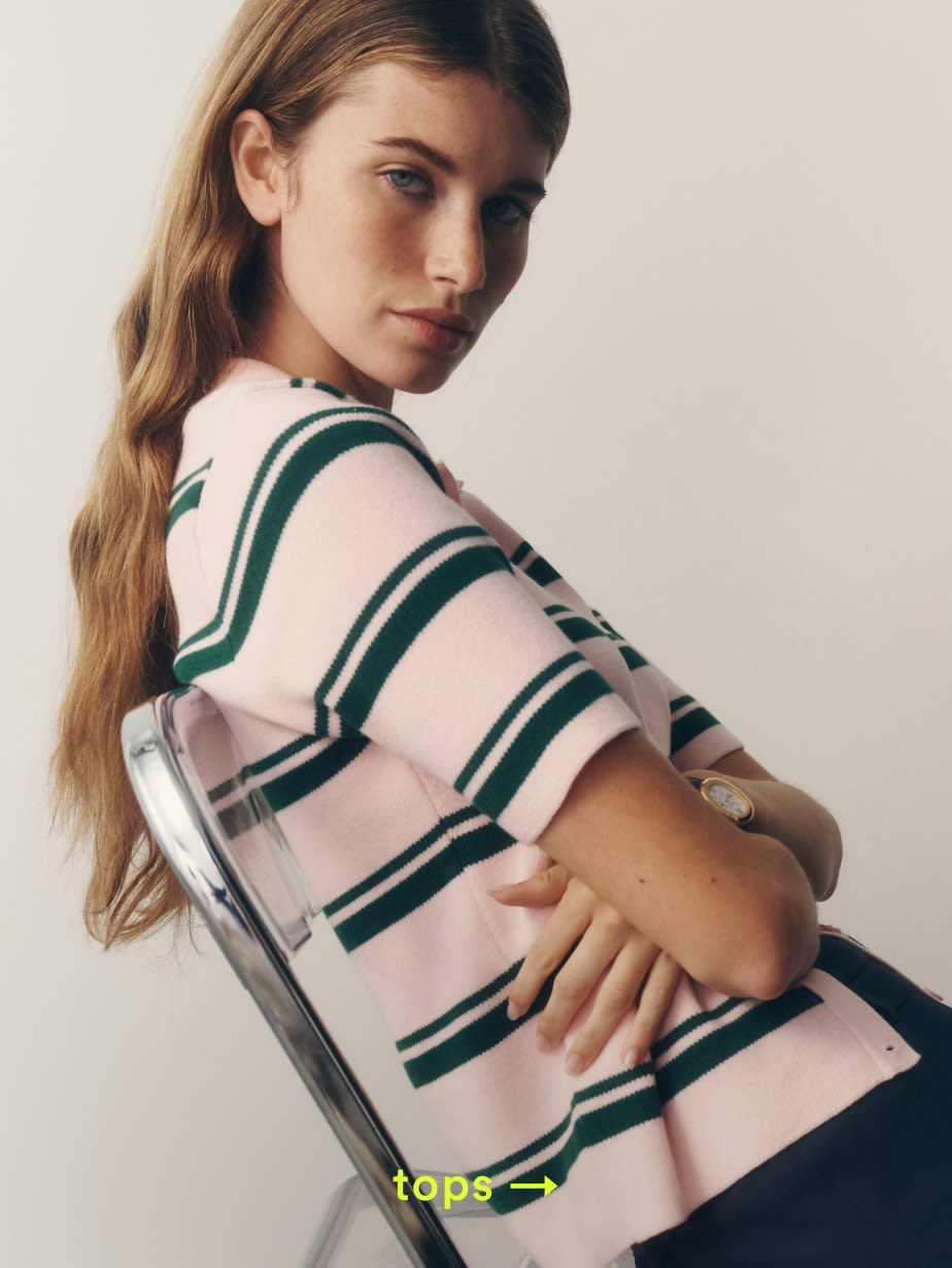 Young woman with long brown hair wearing a pink sweater with green striped pattern, sitting on a chair against beige background. text reads tops.