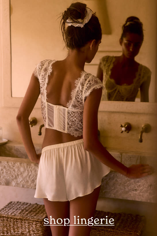 Woman in white lace crop top and shorts standing at bathroom sink, viewed from behind. Text reads "shop lingerie"