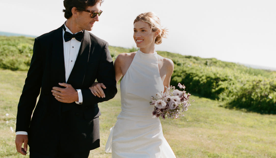 Smiling bride in white halter gown with bouquet walks arm-in-arm with groom in black tuxedo on grassy coastal hillside.