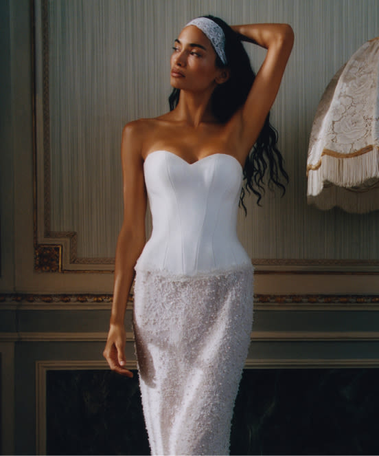 Woman in white strapless corset top and sequined skirt with blue headband, posing elegantly in ornate room with fireplace.
