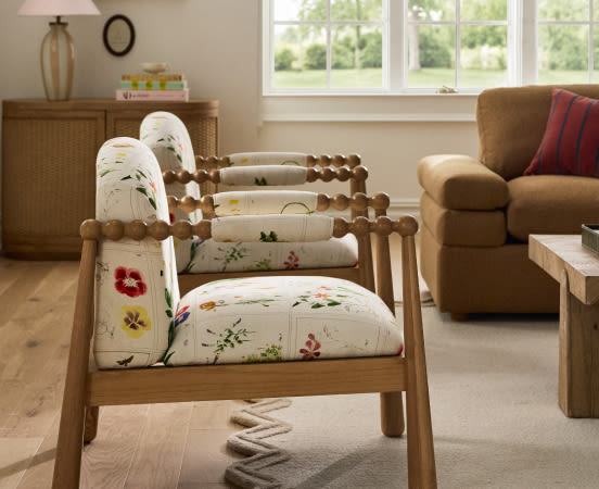 Wooden armchair with white floral embroidered cushions and spindle back in a bright living room with brown furniture and windows.