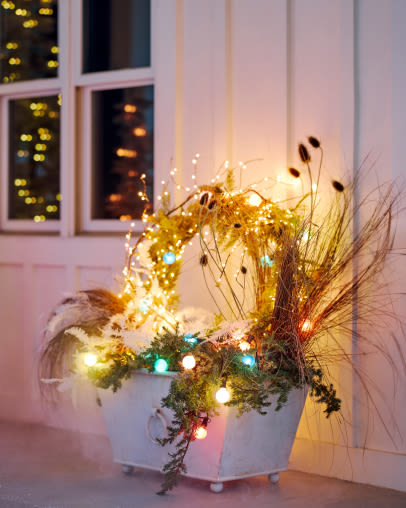 White planter with dried grasses, evergreen branches, and colorful string lights on a porch with windows in background.