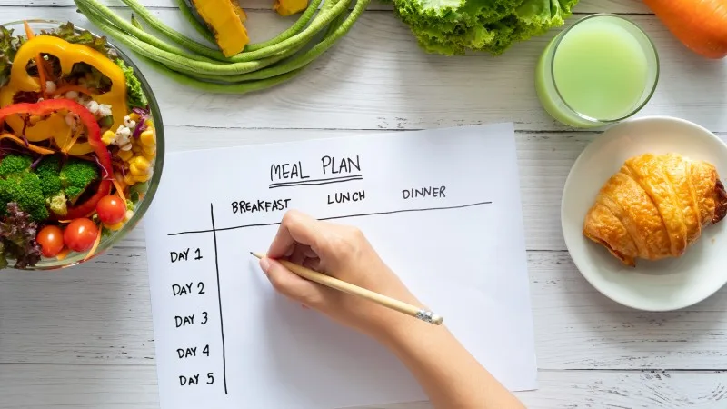 meal plan, food diet and weight loss concept. top view of hand filling meal plan on weekly table with salad and fresh vegetable on dining table