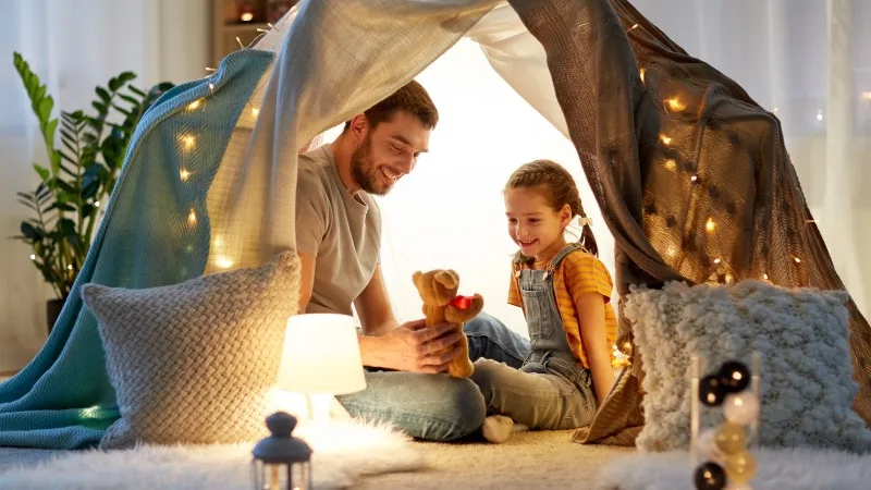 family, hygge and people concept - happy father with teddy bear toy and little daughter playing in kids tent at night at home