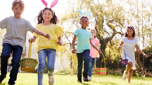 Group Of Children Wearing Bunny Ears Running To Pick Up Chocolate Egg On Easter Egg Hunt In Garden