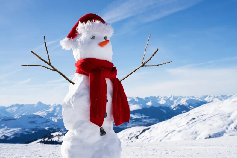 Schneemann mit rotem Schal und Mützen und Stockarmen mit Bergen im Hintergrund.