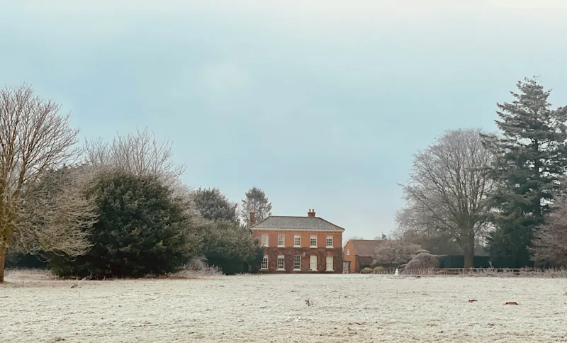 A frost-covered field in winter with a Georgian red-brick manor house in the background, surrounded by bare deciduous trees and evergreen shrubs under a pale grey sky, Sparham, Norfolk.