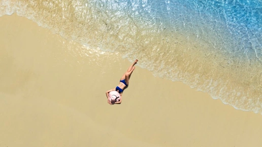 Aerial view of beautiful young woman in black swimsuit lying on sandy beach near blue sea with waves at sunset