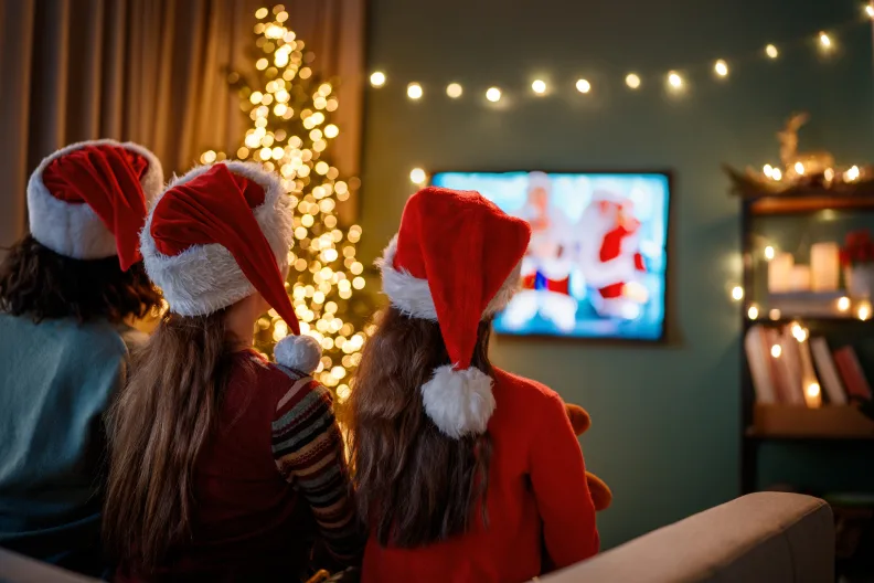 Drei Kinder mit Weihnachtsmützen gucken einen Weihnachtsfilm auf dem Fernseher.