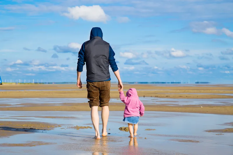 A man and a child walk hand in hand on the beach.