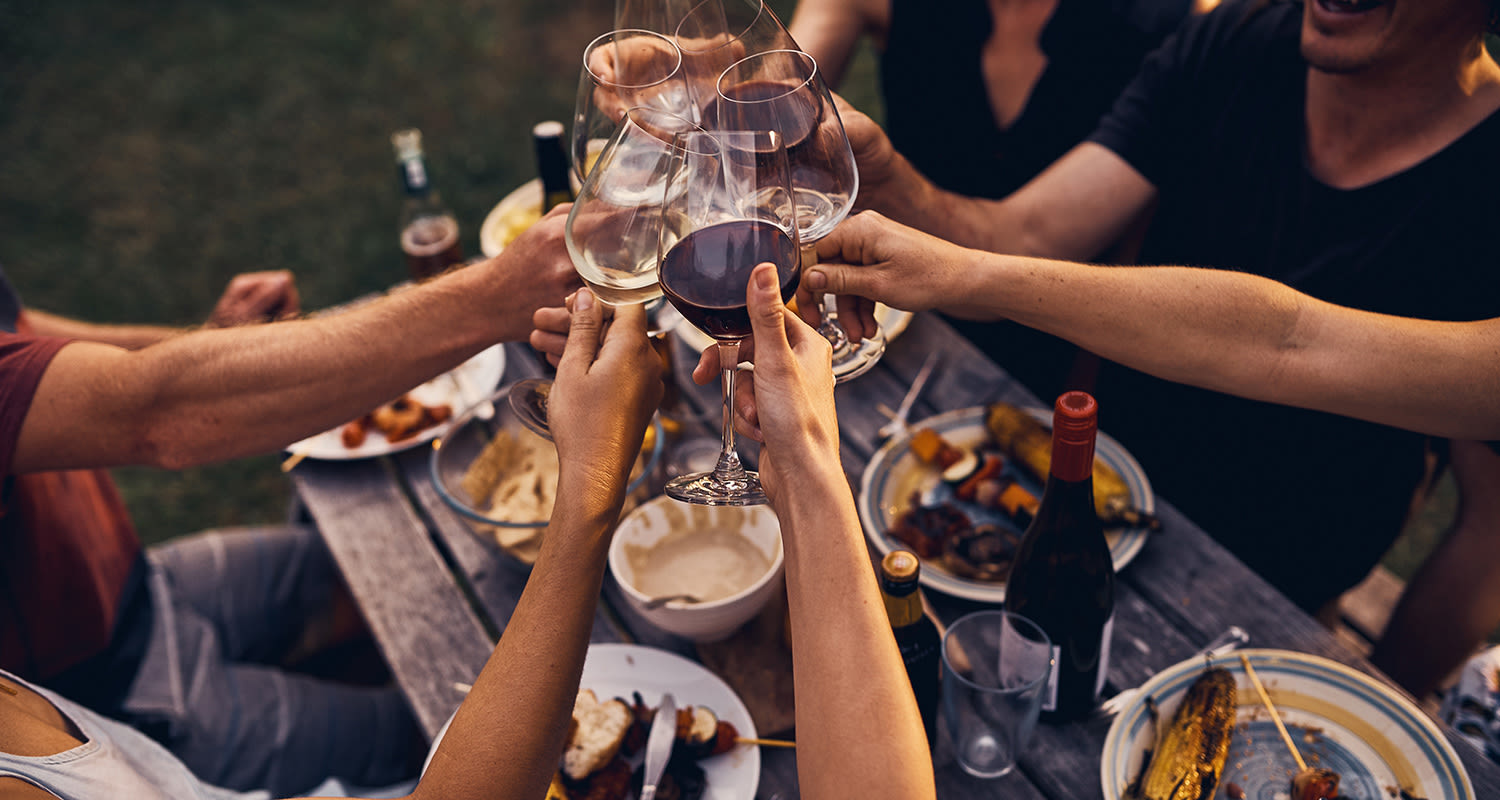 Ihmiset kilistävät viinilasejaan lämpimässä ulkoilmaillallisessa puupöydän ääressä.