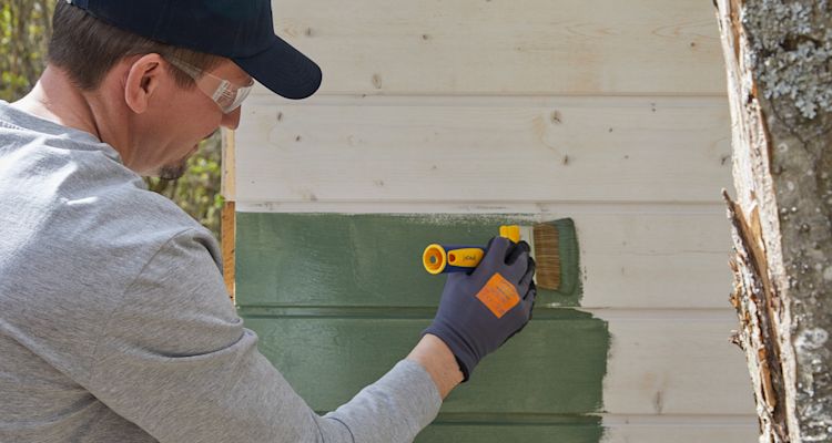 Henkilö maalaa vihreäksi vaaleaa puurakennuksen ulkoseinää tummissa työkäsineissä