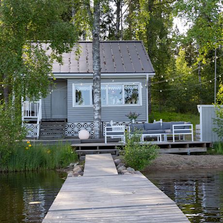 Puisella laiturilla avautuu näkymä harmaalle mökille valkoisine terassikalusteineen.
