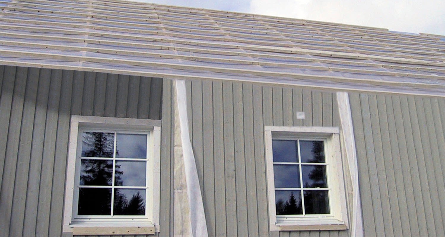 Vihertävä-harmaa puutalo, valkoiset ikkunat heijastavat metsää ja kattolatit näkyvissä.