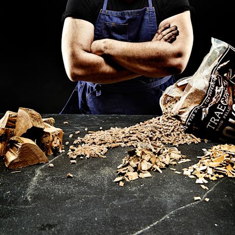 Henkilö sinisessä essussa tummalla taustalla, edessä halkoja, puupellettisäkki ja lastuja.