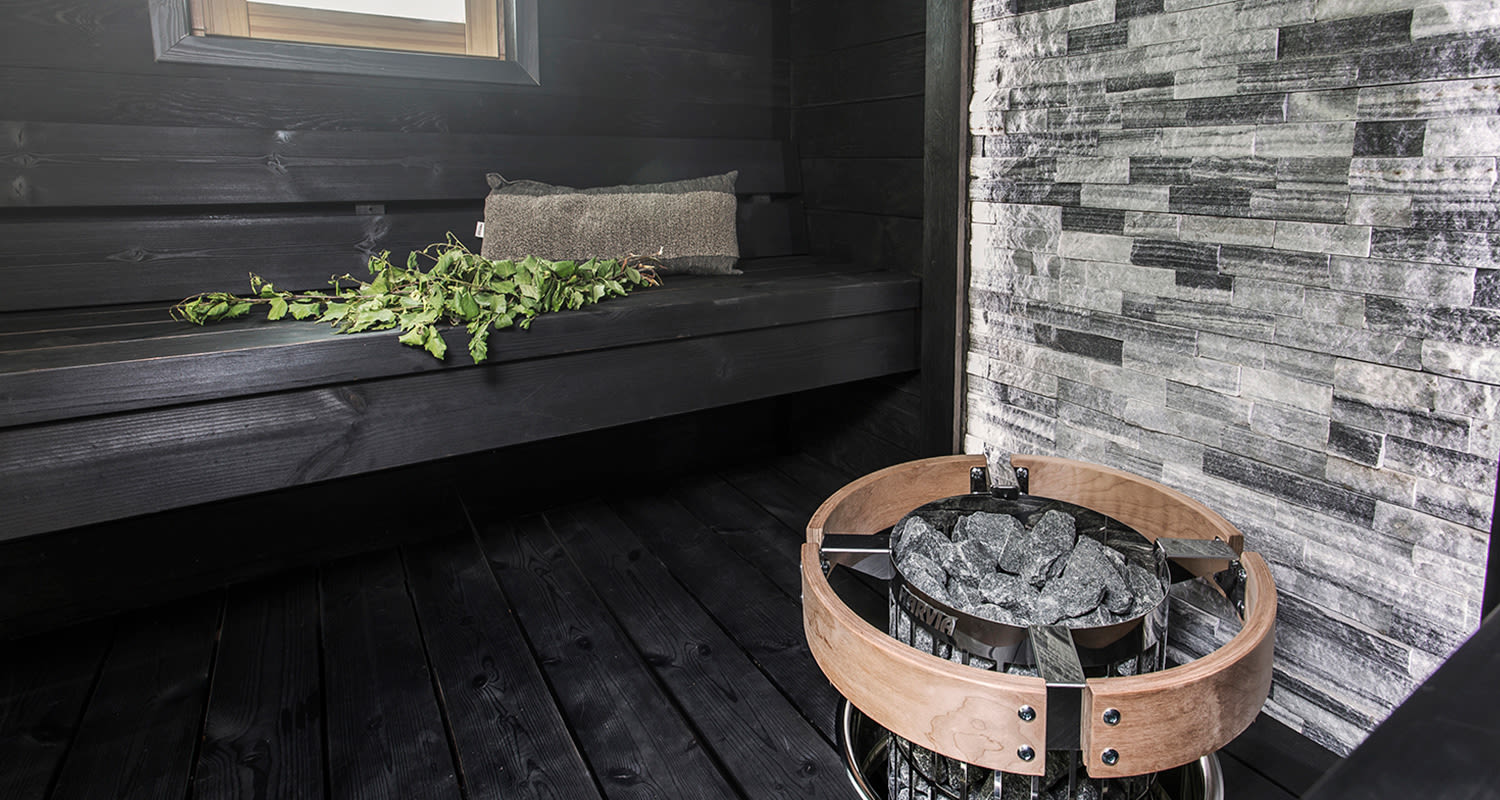 Moderni musta puusauna, harmaa kiviseinä, vihreä vasta penkillä ja kiuas kivineen