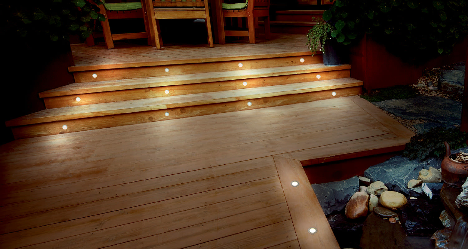 Lämminsävyinen puuterassi kolmiportailla ja upotetuilla valopisteillä kivipihan reunassa