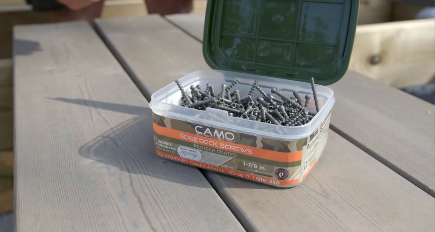 Avoin muovilaatikko oranssi camo-kuvioin täynnä tummia terassiruuveja harmaiden puulautojen päällä