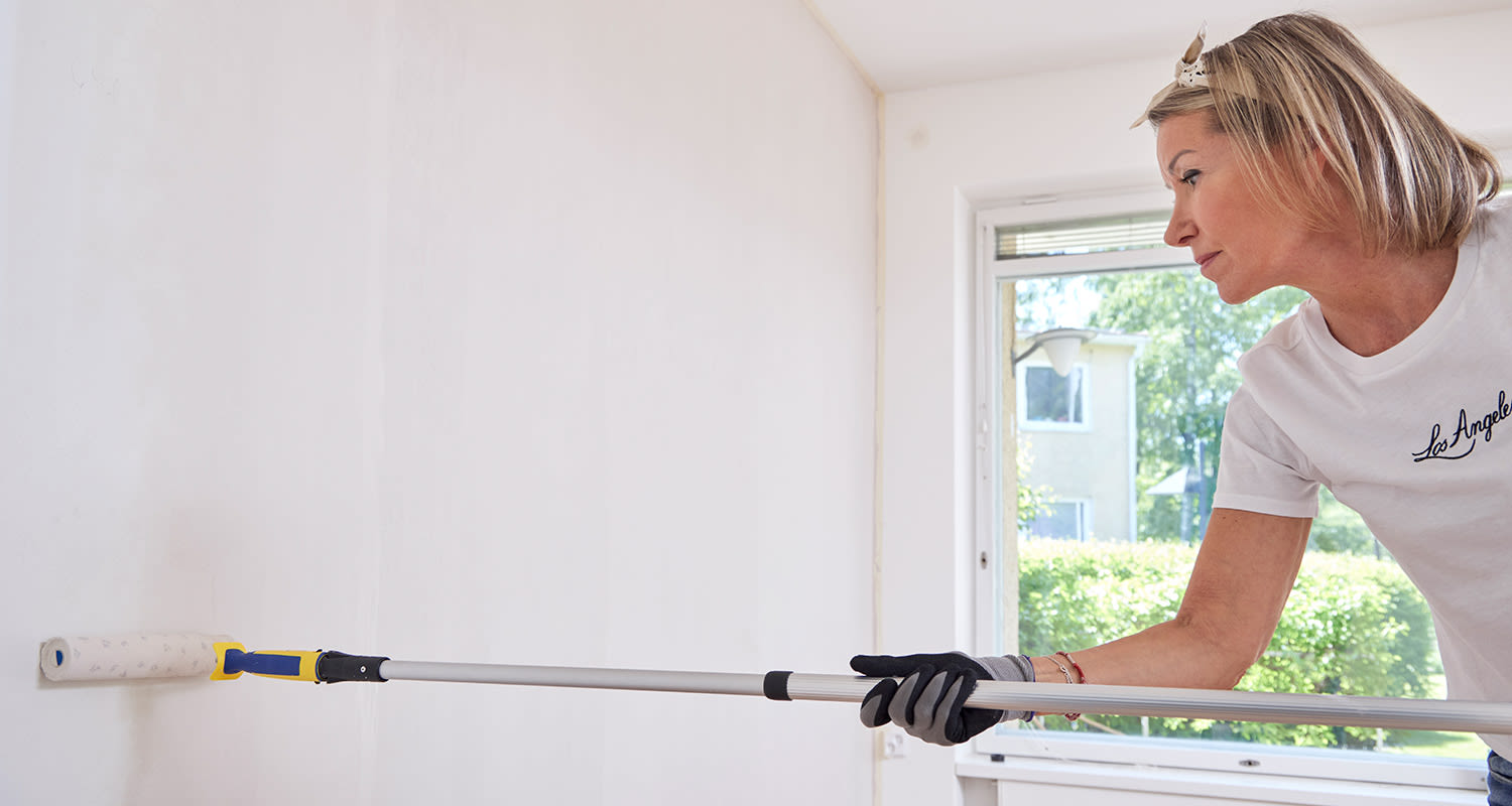 Nainen maalaa sisäseinää vaaleaksi telalla, jossa on jatkovarsi