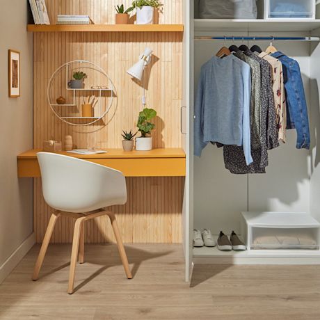 Keltainen pöytä puisella paneeliseinällä, valkoinen tuoli, avoin vaatekaappi ripustetuilla vaatteilla