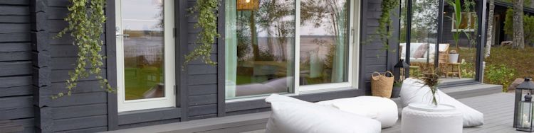 Musta hirsimökki, valkoiset ulkotyynyt terassilla, pikkupöytä maljakolla ja järvinäkyma.