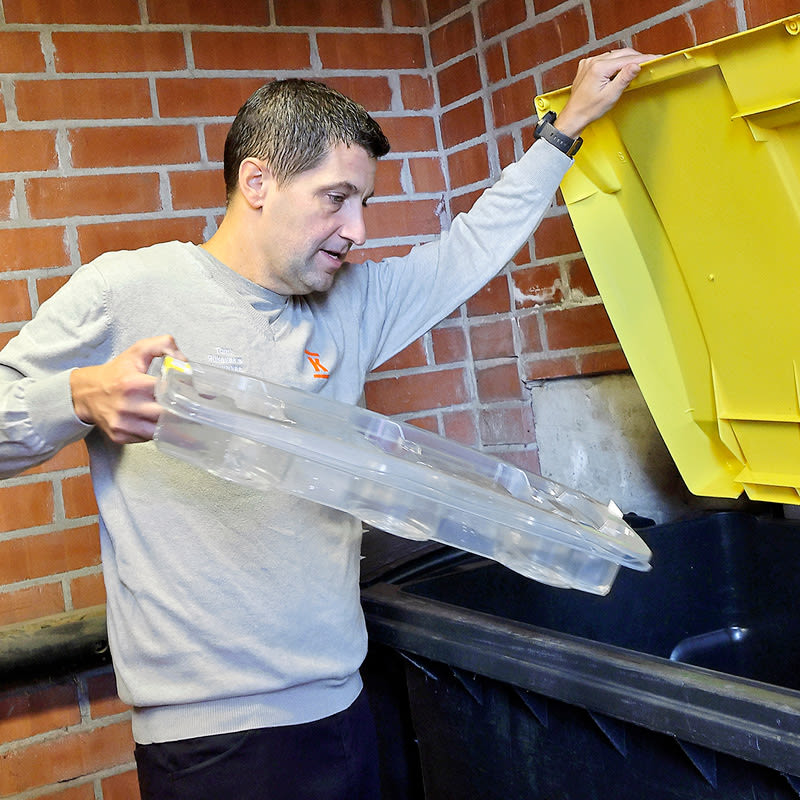 Henkilö laittaa kirkasta muovia keltaiseen kierrätysastiaan tiiliseinän edessä.