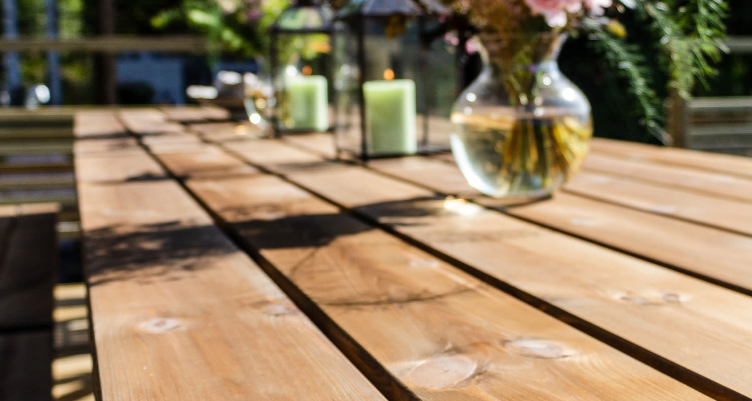 Aurinkoinen puupöytä terassilla, päällä vihreät kynttilät metallilyhdyissä ja kukkamaljakko.