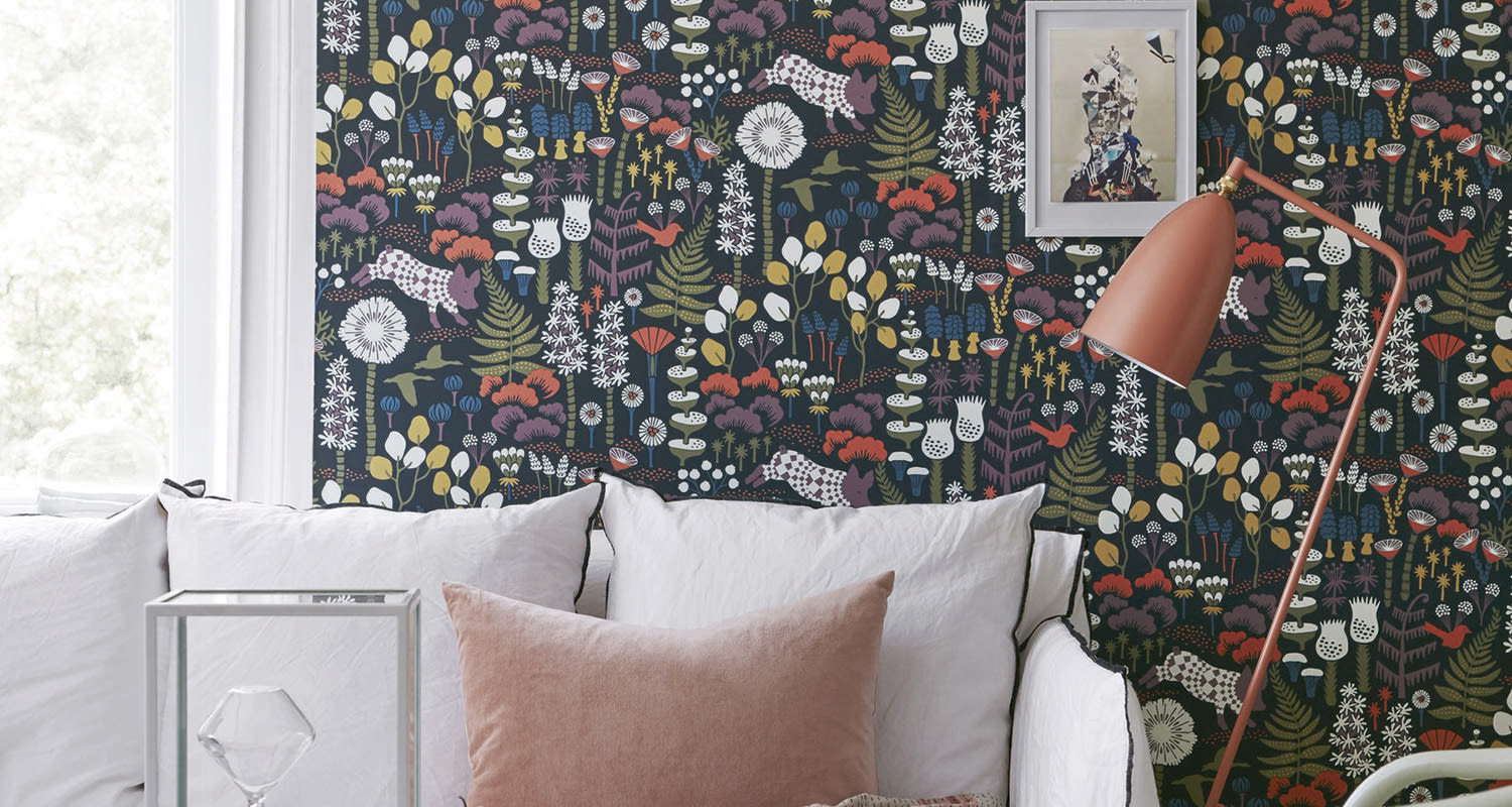 Valkoinen sohva vaaleanpunaisella tyynyllä tummansinistä kukkakuosia vasten ja korallin lattiavalaisin.