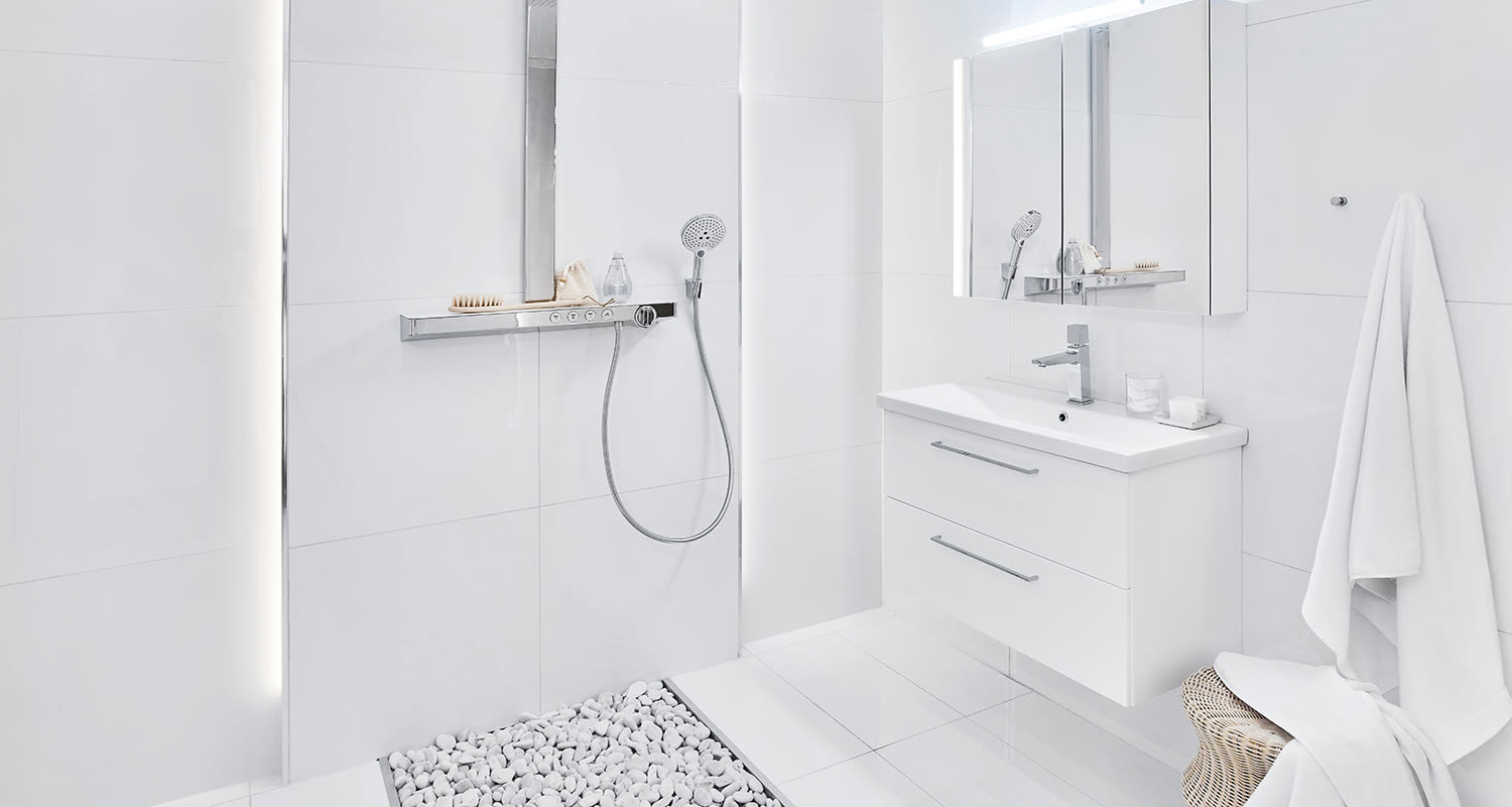 Valkoinen minimalistinen kylpyhuone, kivipohjainen suihku, peili- ja allaskaappi, pyyhkeet.