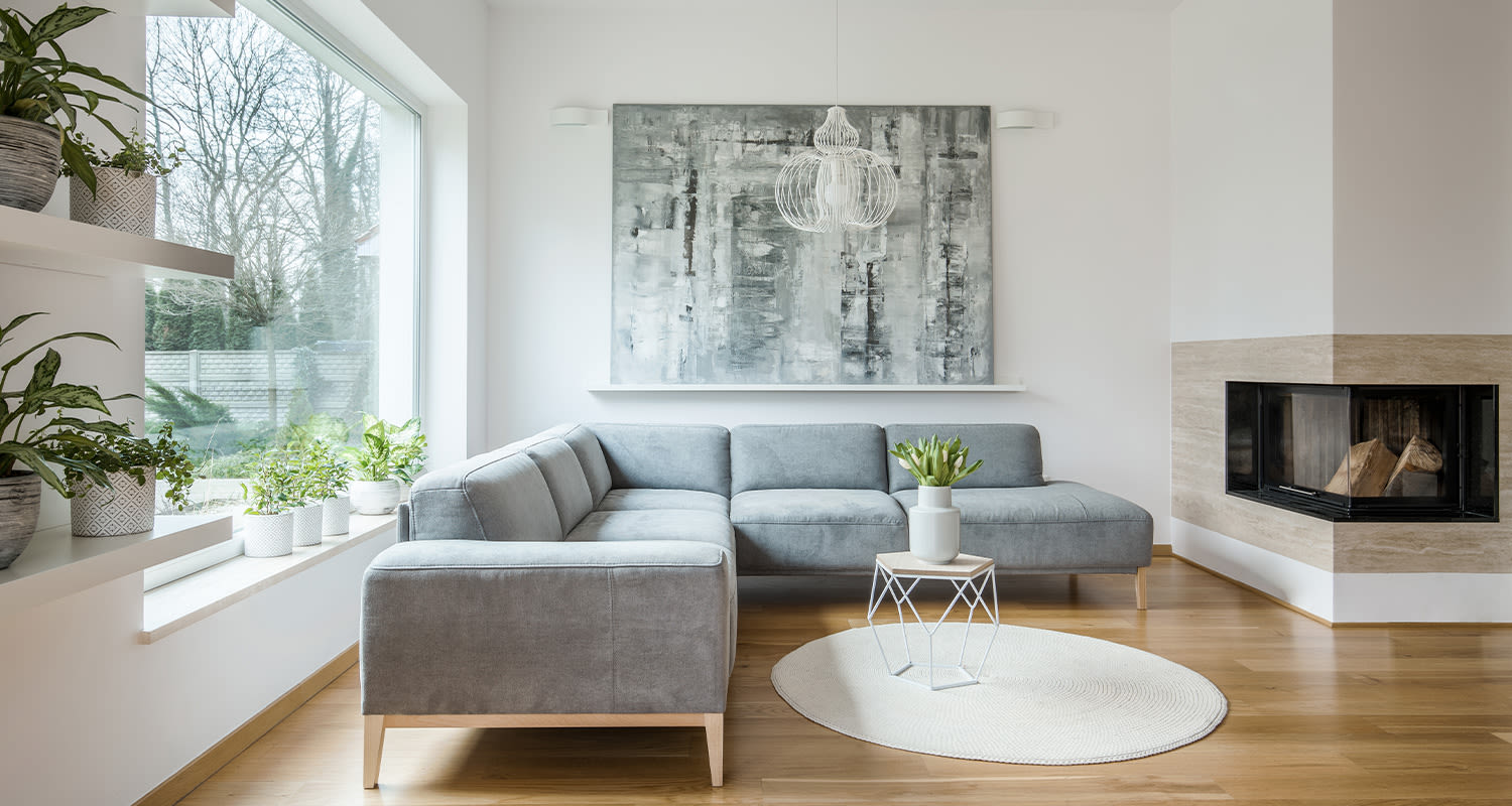 Tyylikäs valoisa olohuone: harmaa kulmasohva, valkoinen takka, puulattia ja vehreät viherkasvit.
