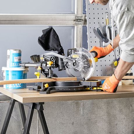 Mies oranssihanskoissa sahaa puulistaa kippisahalla työpajassa, maalipurkit pöydällä.