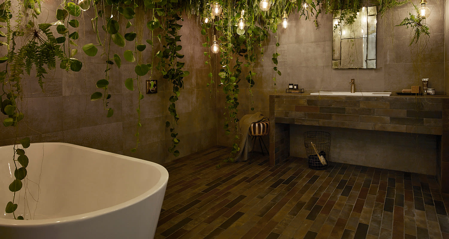 Tumma tiililattia, valkoinen kylpyamme, roikkuvat viherkasvit, peiliallas ja himmeä valaistus.