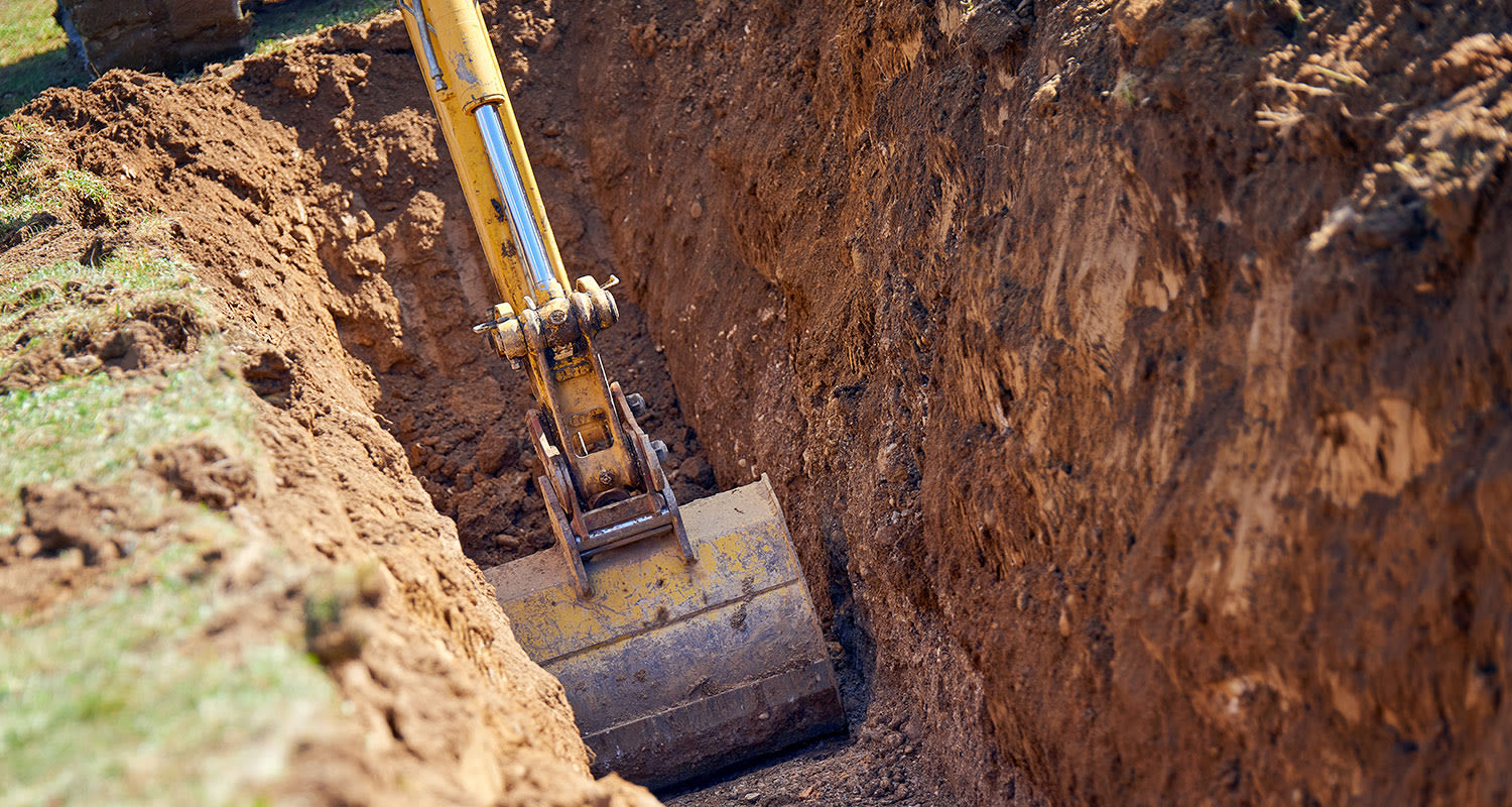 Keltainen kaivinkone kaivamassa syvää ruskeaa kaivantoa, ruohonreunat näkyvissä.