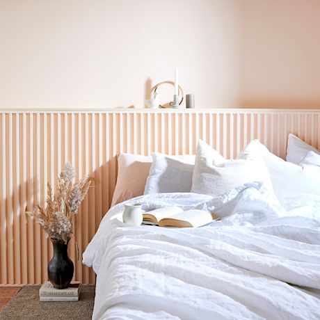 Vaalea sisustus: persikkaseinä, valkoinen sänky avoin kirja ja kuppi, musta kuivaheinämaljakko.