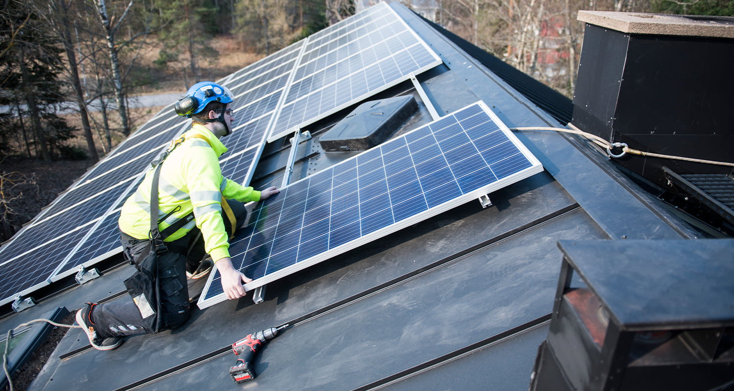 Asentaja keltaisessa takissa ja sinisessä kypärässä kiinnittää aurinkopaneeleita tummalle katolle.