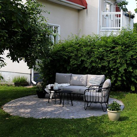 Harmaa ulkosohva kiviterassilla, edessä ruukku, takana vihreä pensas ja valkoinen talo.