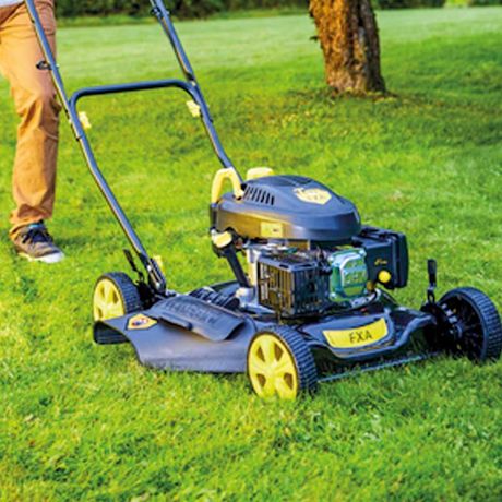 Henkilö työntää mustakeltaista ruohonleikkuria vihreällä nurmikolla puun vieressä.