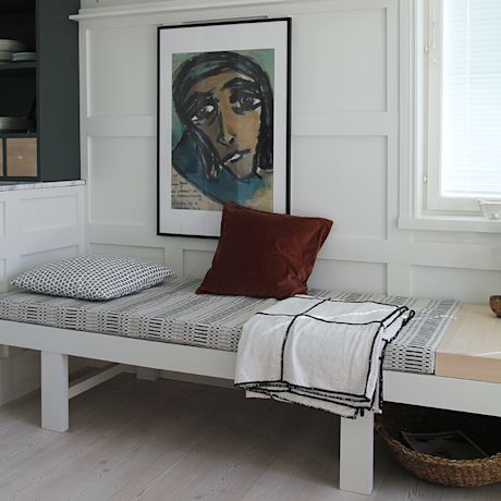 Valkoisella paneeliseinällä penkki mustavalkokuviopatjalla, ruskea tyyny, huopa ja taulu.