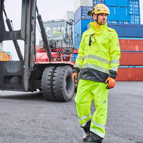 Satamatyöntekijä keltaisessa suojahaalarissa ja kypärässä punaisen trukin ja konttikasojen edessä