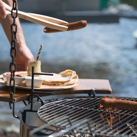 Makkara pihdeissä grillin yllä, leikkuulaudalla tortilloja ja kastikkeita veden äärellä.