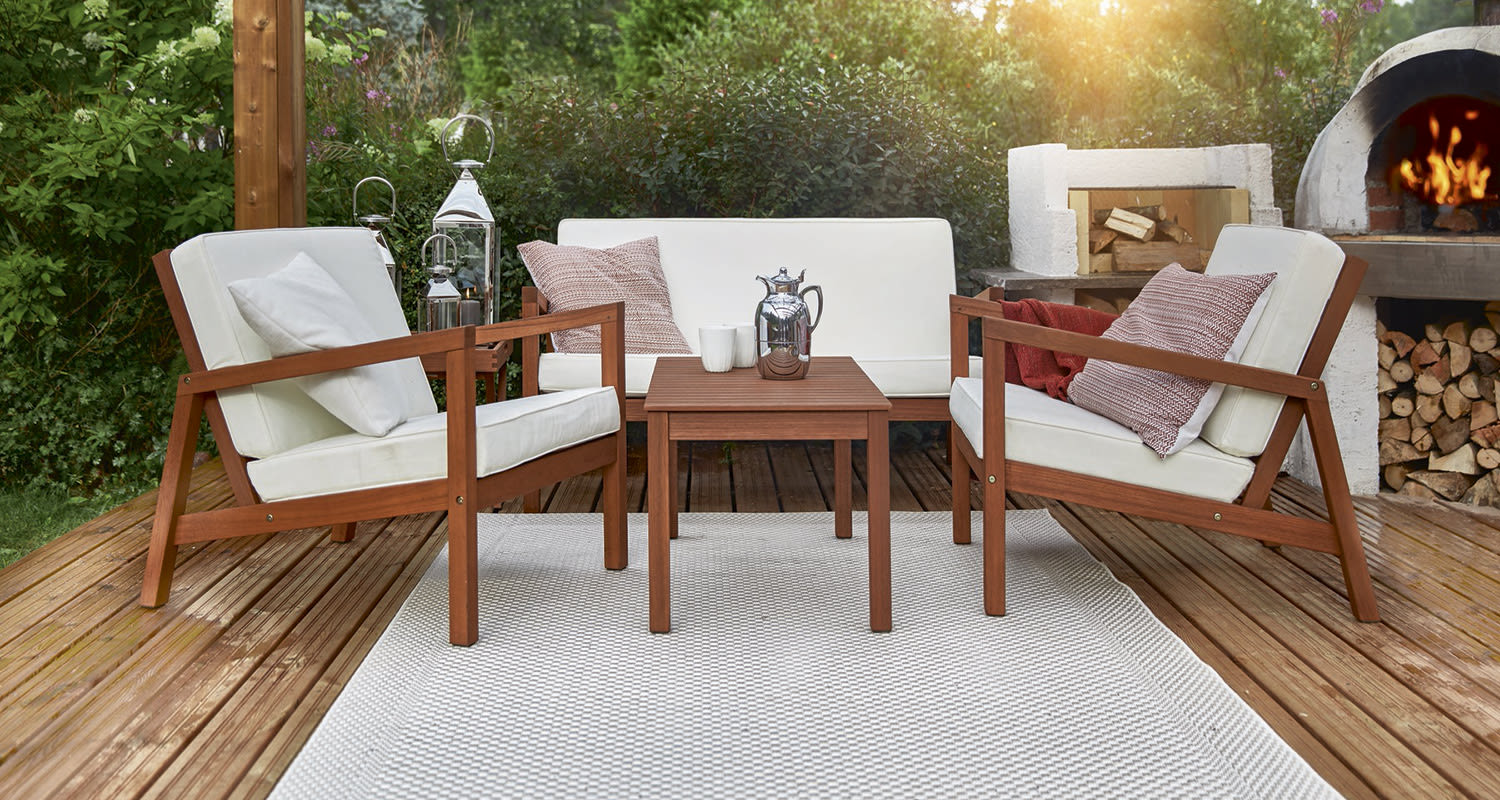 Puinen terassiryhmä valkoisin tyynyin, pikkupöytä hopeateepannulla ja palava puu-uuni