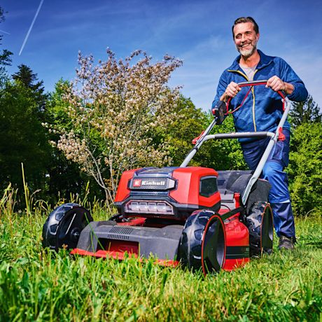 Mies työntää punaista ruohonleikkuria vihreällä nurmikolla aurinkoisessa puutarhassa.