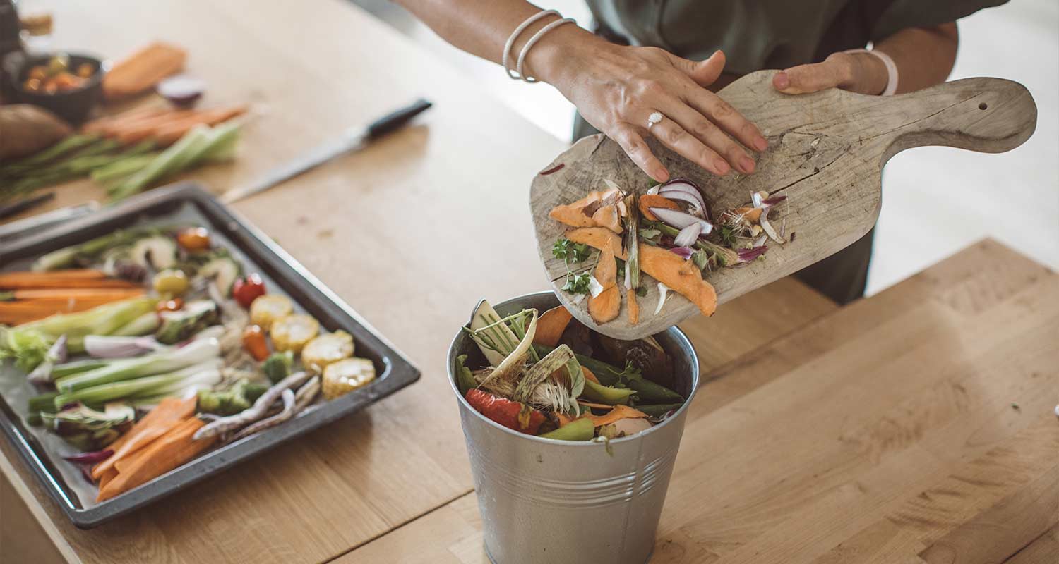 Kaataa porkkanankuoria ja sipulin paloja harmaaseen kompostisankoon, taustalla värikkäitä kasviksia