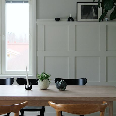 Vaalea tila: tammipöytä, mustat tuolit, pöydällä vaasi vihreällä kasvilla ja kynttilä