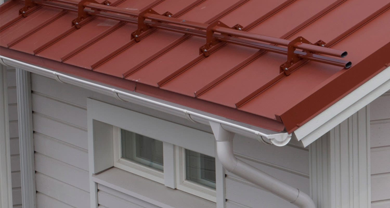 Punainen peltikatto lumikaiteella, valkoinen sadevesikouru, vaalea puupaneeliseinä ja ikkuna